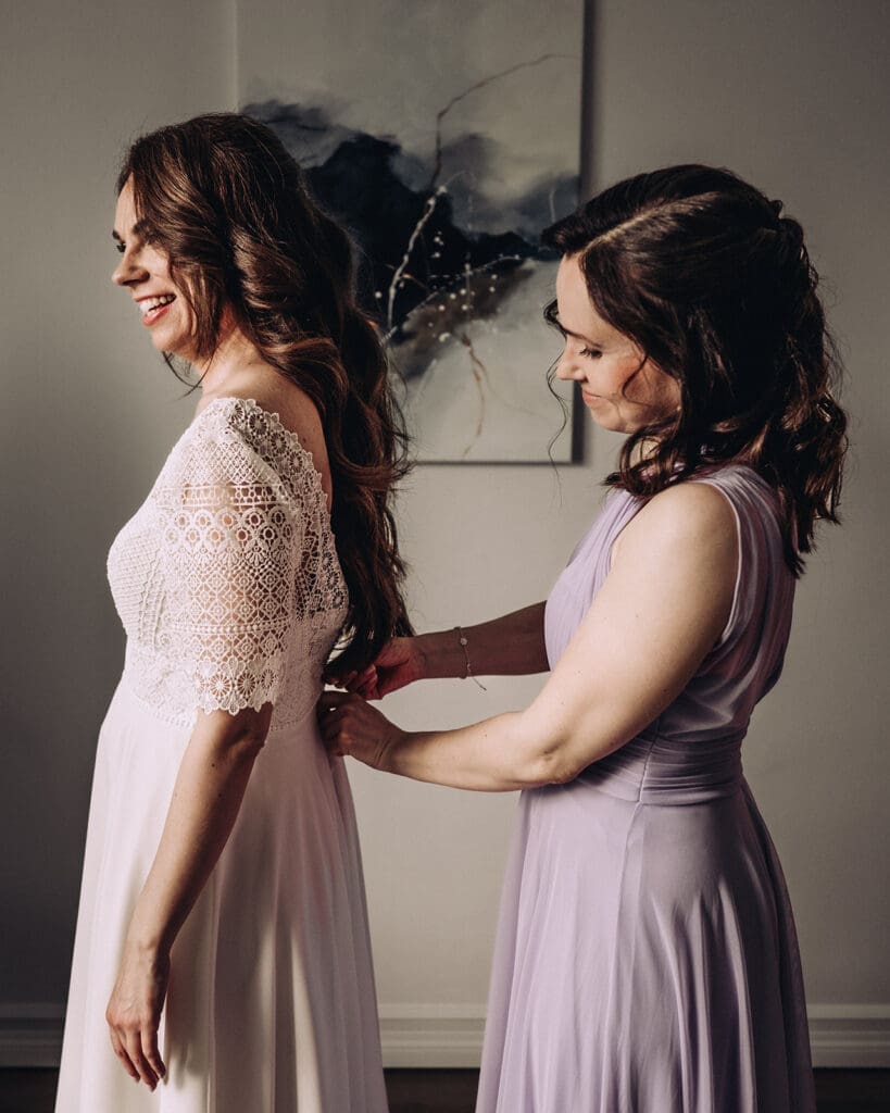 Bridesmaid buttoning bride into lace wedding dress during bridal preparation in Helsinki, candid wedding moment, Miss Moody Photography — Kaaso napittaa morsiamen pitsilistä hääpukuun häävalmisteluissa Helsingissä, aito häähetki, Miss Moody Photography