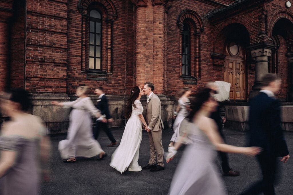 Bride and groom kissing outside Johanneksenkirkko church in Helsinki, wedding guests moving around them, Miss Moody Photography — Morsian ja sulhanen suutelevat Johanneksenkirkon edustalla Helsingissä, häävieraat liikkuvat heidän ympärillään, Miss Moody Photography