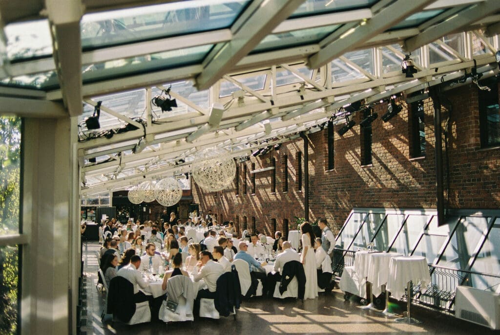 Ravintola Sipuli wedding reception interior Helsinki, industrial glass ceiling venue with wedding guests, film photography, Miss Moody Photography — Ravintola Sipulin häävastaanoton sisätila Helsingissä, teollinen lasikattovenue häävierailla, filmivalokuvaus, Miss Moody Photography
