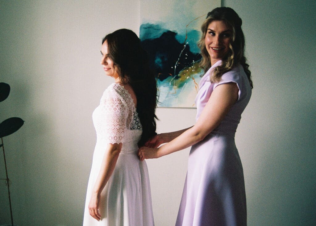Bridesmaid helping bride into wedding dress during bridal prep in Helsinki, Miss Moody Photography — Kaaso auttaa morsianta pukeutumaan hääpukuun häävalmisteluissa Helsingissä, Miss Moody Photography