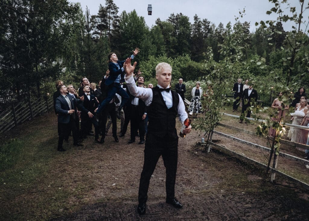 Groom throwing garter to wedding guests outdoors at Aurantola manor wedding in Jaala Kymenlaakso Finland, guests jumping to catch it, moody documentary wedding photography by Miss Moody Photography | Sulhanen heittää sukkanauhaa häävieraille ulkona Aurantolan kartanohäissä Jaalassa Kymenlaaksossa, vieraat hyppäävät kiinni ottaakseen sen, hääkuvaus Miss Moody Photography