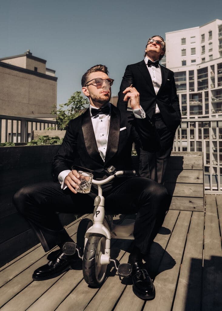 Groom and his bestman on rooftop terrace in Pasila Helsinki, one sitting on toddler tricycle holding whisky glass in tuxedo, editorial wedding photography, Miss Moody Photography — Sulhanen ja bestman kattoterassilla Pasilassa Helsingissä, yksi istuu lasten kolmipyörällä pitäen viskiä smokingissa, editoraalinen hääkuvaus, Miss Moody Photography