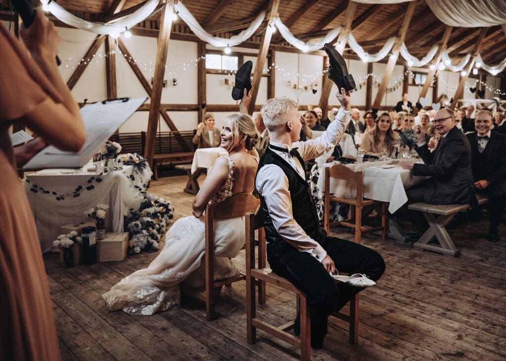 Bride and groom playing the shoe game at Aurantola wedding reception in rustic barn venue Jaala Kymenlaakso Finland, guests laughing in background, documentary wedding photography by Miss Moody Photography | Morsian ja sulhanen kenkäpelissä Aurantolan häävastaanottossa rustiikkisessa latotilassa Jaalassa Kymenlaaksossa, vieraat nauravat taustalla, hääkuvaus Miss Moody Photography
