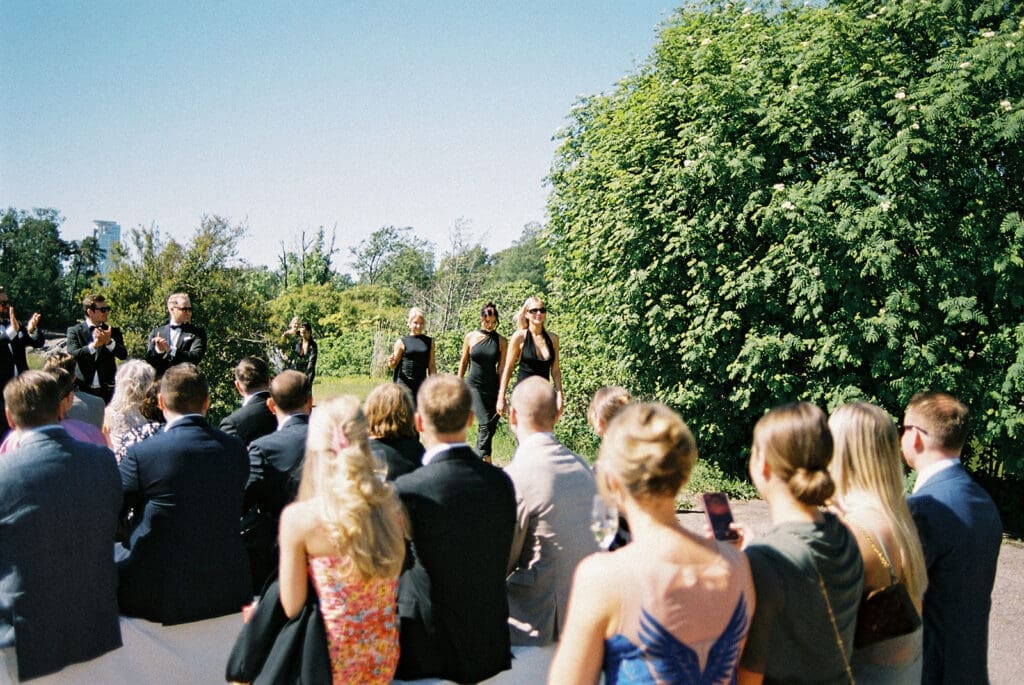 Bridesmaids in black dresses walking to outdoor wedding ceremony at Kulosaari Casino Helsinki, guests watching, film photography, Miss Moody Photography — Kaasot mustissa mekoissa kävelevät ulkohäihin Kulosaaren Casinolla Helsingissä, vieraat katsovat, filmivalokuvaus, Miss Moody Photography