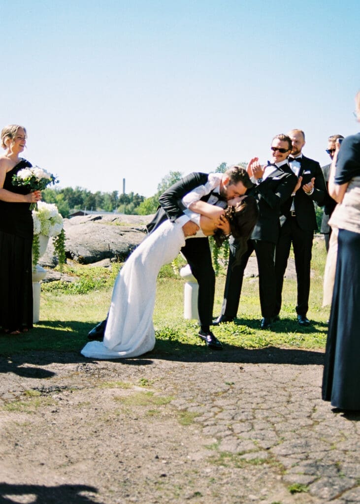 Bride and groom first kiss dip on Helsinki rocks at Kulosaari Casino outdoor ceremony, wedding party celebrating around them, film photography, Miss Moody Photography — Morsian ja sulhasen ensimmäinen suudelma kalliolla Kulosaaren Casinon ulkoseremoniassa Helsingissä, hääseurue juhlistaa ympärillä, filmivalokuvaus, Miss Moody Photography
