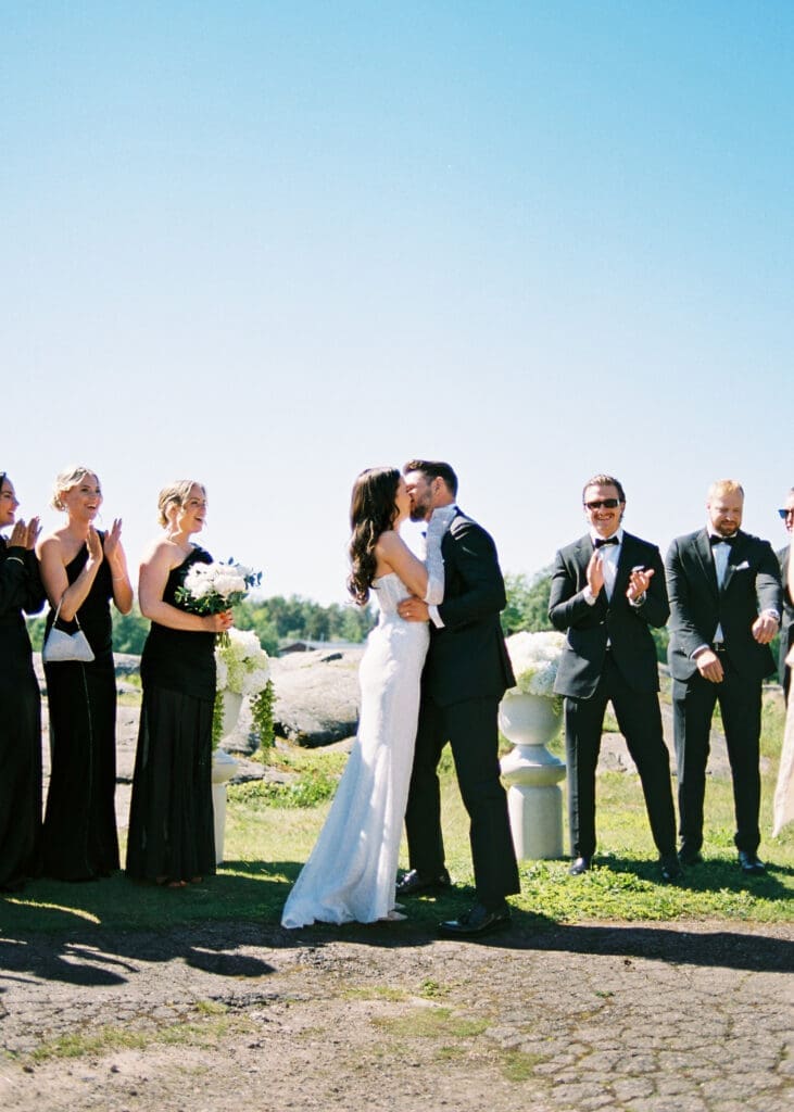 Bride and groom first kiss at outdoor wedding ceremony on Helsinki rocks Kulosaari Casino, all black wedding party celebrating, film photography, Miss Moody Photography — Morsiamen ja sulhasen ensimmäinen suudelma ulkohäissä Helsingin kalliolla Kulosaaren Casino, koko musta hääseurue juhlistaa, filmivalokuvaus, Miss Moody Photography