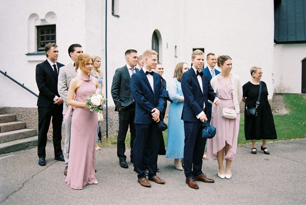 Wedding guests gathered outside Valkeala kirkko white church after ceremony in Finland, 35mm film wedding photography by Miss Moody Photography Kymenlaakso | Häävieraat Valkealan kirkon ulkopuolella vihkimisen jälkeen, filmikuvaus hääkuvaaja Miss Moody Photography Kymenlaakso