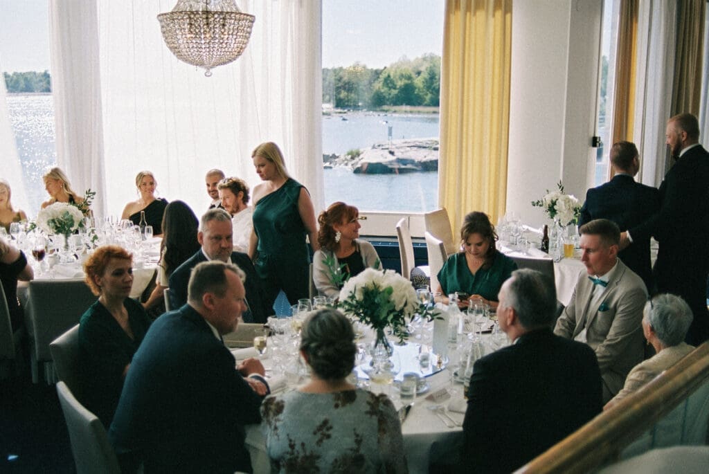 Wedding reception at Kulosaari Casino Helsinki with waterfront view through windows, guests seated at tables, film photography, Miss Moody Photography — Häävastaaanotto Kulosaaren Casinolla Helsingissä vesistönäkymällä ikkunoiden läpi, vieraat istuvat pöydissä, filmivalokuvaus, Miss Moody Photography