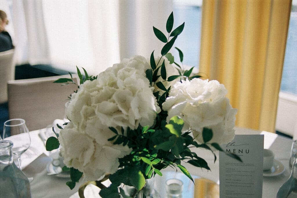 Wedding reception table detail with white hydrangea centerpiece and menu card at Kulosaari Casino Helsinki, film photography, Miss Moody Photography — Häävastaanoton pöytäyksityiskohta valkoisella hortensiakeskikappaleella ja menukorttilla Kulosaaren Casinolla Helsingissä, filmivalokuvaus, Miss Moody Photography