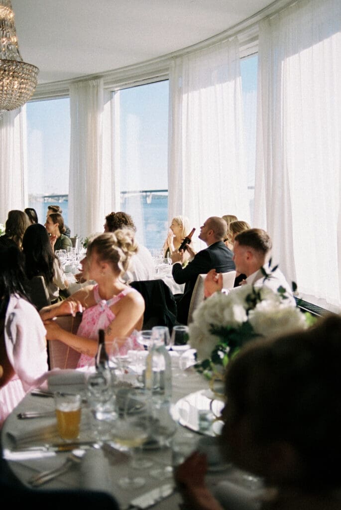 Wedding reception guests seated at Kulosaari Casino Helsinki with panoramic waterfront windows and crystal chandelier, film photography, Miss Moody Photography — Häävieraat istuvat Kulosaaren Casinolla Helsingissä panoraamavesistöikkunoiden ja kristallikruunun kanssa, filmivalokuvaus, Miss Moody Photography