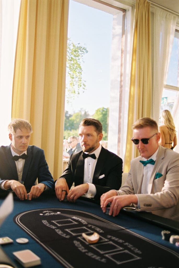 Groomsmen in tuxedos playing poker at wedding reception Kulosaari Casino Helsinki, sunglasses and bow ties, yellow curtains and waterfront windows, film photography, Miss Moody Photography — Sulhasen kaverit smokingeissa pelaavat pokeria häävastaanottolla Kulosaaren Casinolla Helsingissä, aurinkolasit ja rusettikravatit, keltaiset verhot ja vesistöikkunat, filmivalokuvaus, Miss Moody Photography