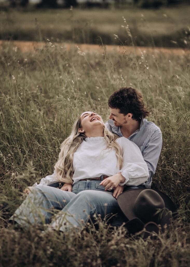 Woman laughing with her head tipped back as her fiancé smiles at her in long grass