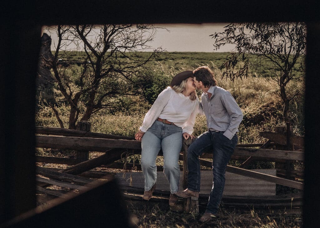 Couple kissing while sitting on a timber fence with gum trees behind them